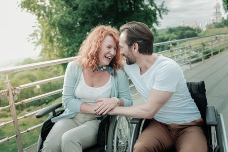 Lets walk. Smiling red haired female laughing at funny joke and embracing her man while sitting on the wheelchairの写真素材