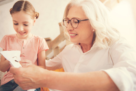 Happy positive elderly woman smiling and embroidering while spending time with her granddaughter