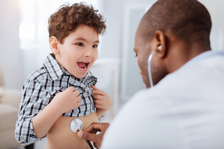 Cold stethoscope. Positive vigorous boy opening mouth while male doctor using stethoscope and examining himの写真素材