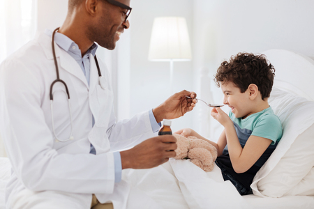 Cough medicine. Joyful merry boy laughing while male doctor looking at him and stretching spoon with medicationの写真素材