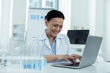 Great results. Pretty exuberant dark-haired researcher smiling and wearing a uniform while working on her laptop in the labの写真素材