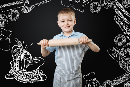 Tasty bakery. Cheerful active little boy smiling and feeling ready to start cooking while standing with a big rolling pin in his handsの写真素材