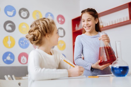 Pleasant conversation. Charming teenage girl talking to her friend during the chemistry class while holding a big flask with a red liquidの写真素材
