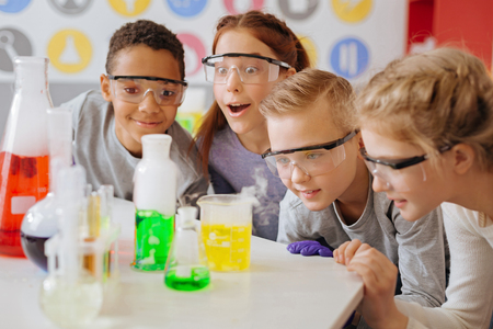Fond of chemistry. The group of upbeat teenage students being gathered around the table in their school lab and observing the final stage of their chemical experiment with excitementの写真素材