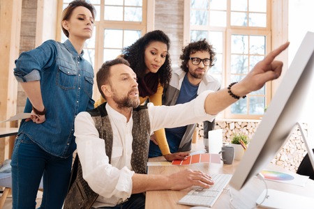Look here. Good-looking concentrated bearded man working on his computer and showing something on the screen to his colleagues while sitting at the tableの写真素材