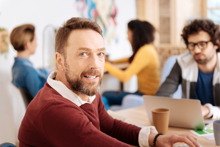 My office. Good-looking alert bearded man smiling and sitting at the table while his colleagues working in the backgroundの写真素材
