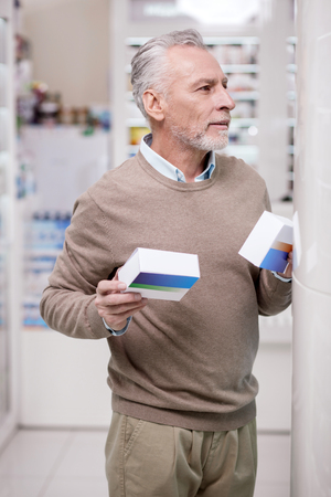 Several variants. Pensive senior man standing in drugstore while holding medicationsの写真素材