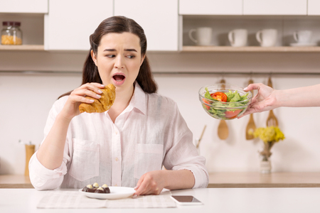 Tasty not healthy. Unsettled nice woman staring at salad while consuming croissantの写真素材