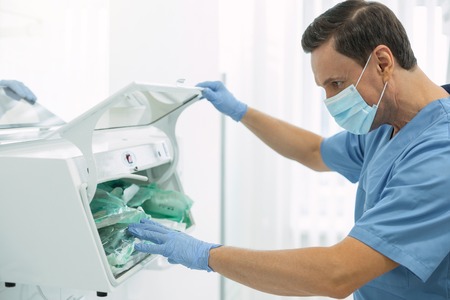 My routine work. Serious dark-haired dentist wearing a uniform and looking for instrumentsの写真素材