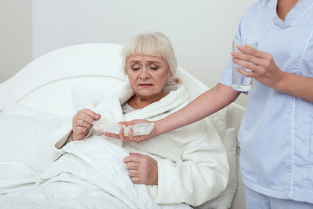Life with drugs. Dismal elder woman lying in bed while consuming drugsの写真素材