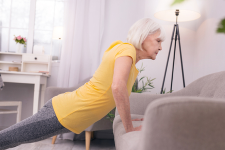 Strong woman. Athletic senior woman resting her hands on the sofa and doing push-ups during her morning workoutの写真素材