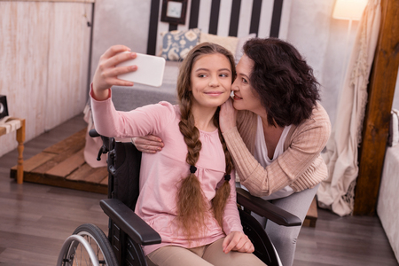 Charming photo. Satisfied crippled girl using phone while woman kissing herの写真素材