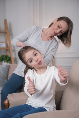 Cheerfulness. Delighted nice girl sitting on the sofa and her mom combing her hairの写真素材