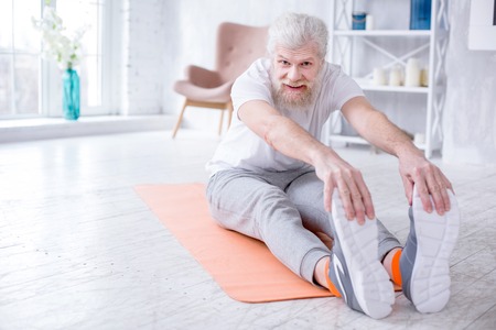 Supple body. Upbeat senior man bending forward and touching his toes while sitting on the yoga mat and smiling at the cameraの写真素材