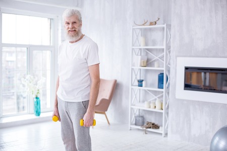 Sporty elderly. Pleasant senior man standing in the middle of the living room and posing for the camera while holding dumbbellsの写真素材