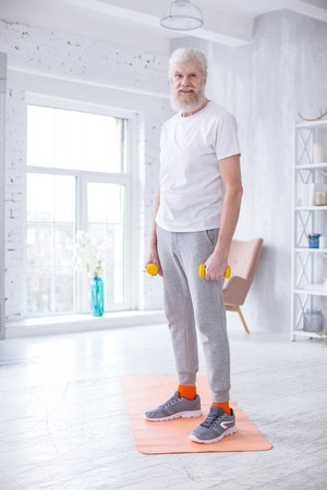 In good shape. Handsome elderly man standing on the yoga mat and holding dumbbells while smiling at the cameraの写真素材
