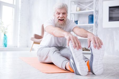 Stretching muscles. Pleasant elderly man sitting on the yoga mat, tugging at his toes and smiling widely, being happy about his supplenessの写真素材