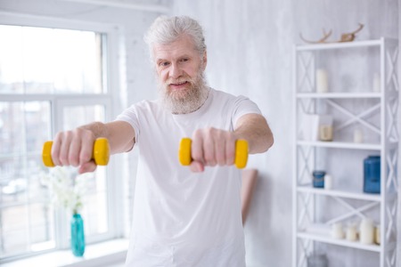 Strengthening his arms. Pleasant elderly man in a white t-shirt working out with dumbbells while standing in the living roomの写真素材