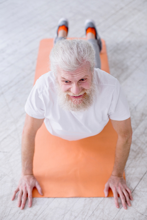 Yoga enthusiast. The top view of a handsome elderly man smiling pleasantly while standing in upward-facing dog pose on a yoga matの写真素材