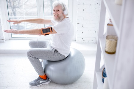 Mood-boosting workout. The top view of a pleasant elderly man smiling at the camera while working out on the fitness ball and listening to music in headphonesの写真素材