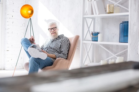 Advanced user. Pleasant senior man sitting in an armchair and smiling at the camera while using a tablet and reading something from itの写真素材