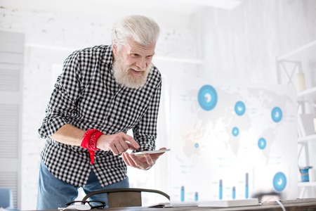 Innovative technologies. Charming elderly man studying the chart on the transparent screen and searching the data from on his phoneの写真素材