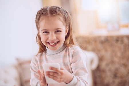 Modern smartphone. Happy delighted girl holding her smartphone while using it for entertainmentの写真素材