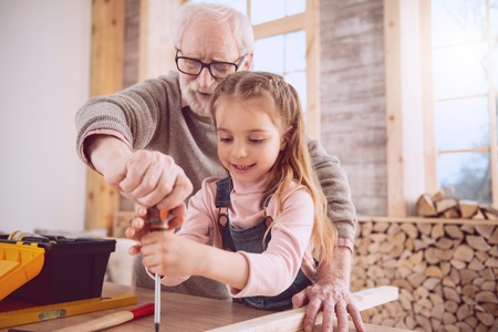 Positive cheerful man holding a screwdriver while working together with his granddaughter in the workshopの写真素材