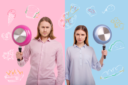 Serious bloggers. Calm serious experienced bloggers holding colorful frying pans and frowning while posing for a photoの写真素材