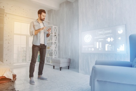 Serious blogger. Concentrated experienced blogger standing with a modern tablet in his hands and looking attentively at the big screen on the wallの写真素材