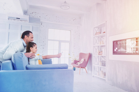 Watching TV. Cheerful smiling man leaning to his daughter while watching TV with herの写真素材