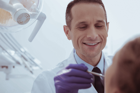Teeth examination. Appealing male dentist grinning while treating patients teethの写真素材