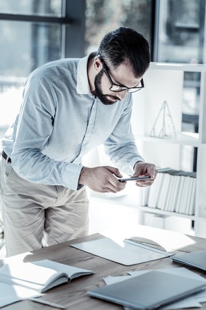 Make a photo. Busy serious unshaken man standing in the office holding cellphone and tilting under the table.の写真素材