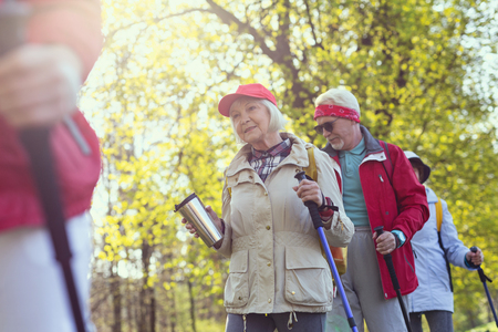 Sunny day. Delighted aged woman holding a thermos bottle while hiking with her friendsの写真素材