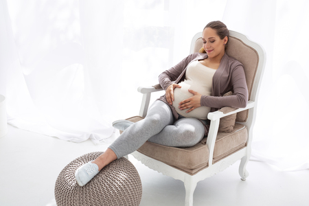 Baby in tummy. Pleased pregnant woman resting in chair and caressing tummyの写真素材