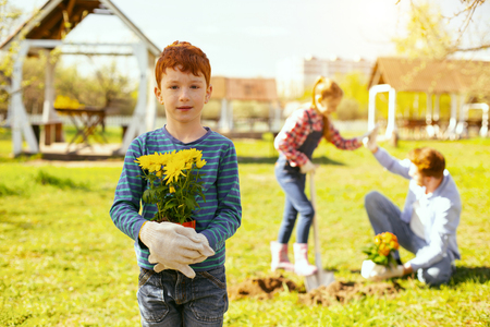 Positive mood. Cute young boy looking at you while standing in his family garden with a flower potの写真素材