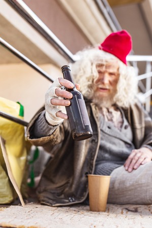 Alcohol addicted. Selective focus of an empty bottle being in hands of a sad homeless manの写真素材