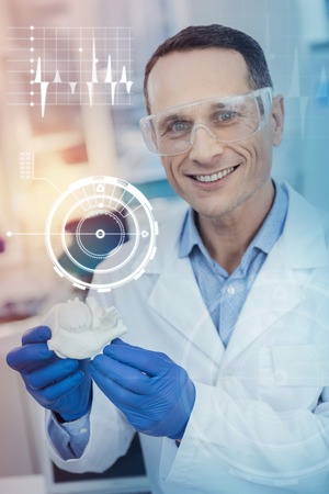 Showing heart miniature. Positive smiling doctor looking happy and showing a little realistic model of a human heartの写真素材