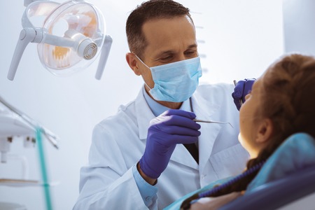 Sterile workplace. Competent dentist wearing mask on face while treating his little patientの写真素材