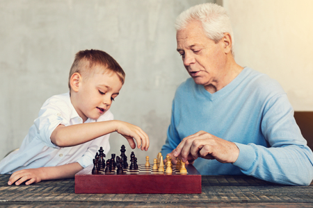 Playing chess. Attentive smart serious men of different generations playing chess while sitting at the tableの写真素材