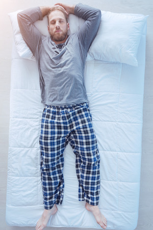 Taking a break. Top view on a millennial man lying in bed with his hands over the head while falling asleep alone.の写真素材