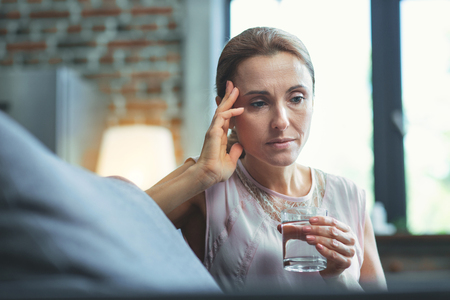 Severe symptoms. Low angle of sad mature woman carrying glass of water and dabbing headの写真素材