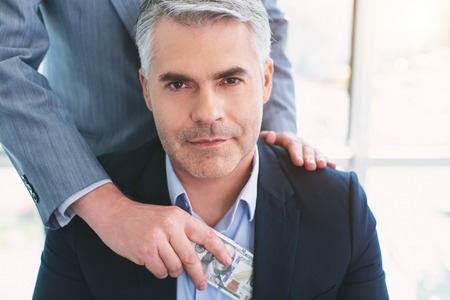 Delighted. Content grey-haired businessman smiling and his colleague putting money into his pocketの写真素材