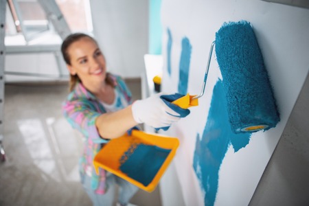 Joyful dark-haired woman holding a roller and painting the walls