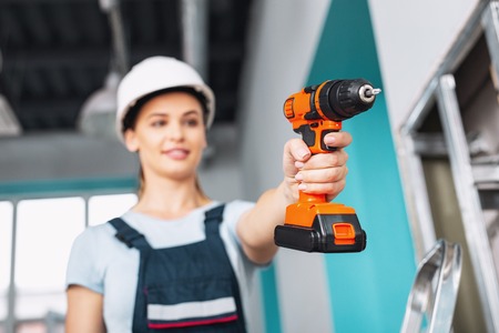 Inspired experienced builder wearing a uniform and working with a drill