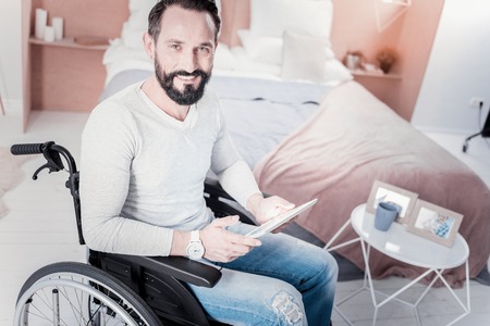 Good mood. Cheerful handicapped man sitting in a wheelchair and holding a tabletの写真素材