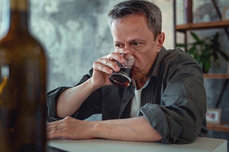 Drinking cognac. Mature grey-haired man wearing dark shirt drinking glass of cognac after some...