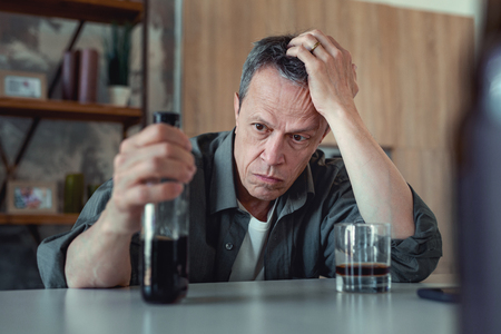 Bottle of cognac. Dark-eyed mature man looking at the bottle of cognac after stressful events sitting in the kitchenの写真素材
