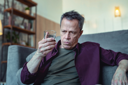 Depressed man. Depressed mature grey-haired man looking at his glass of vodka while sitting on the floor near sofaの写真素材