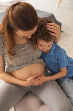 Curious boy. Handsome cute curly haired cute boy feeling rather curious while touching pregnant belly of his motherの写真素材